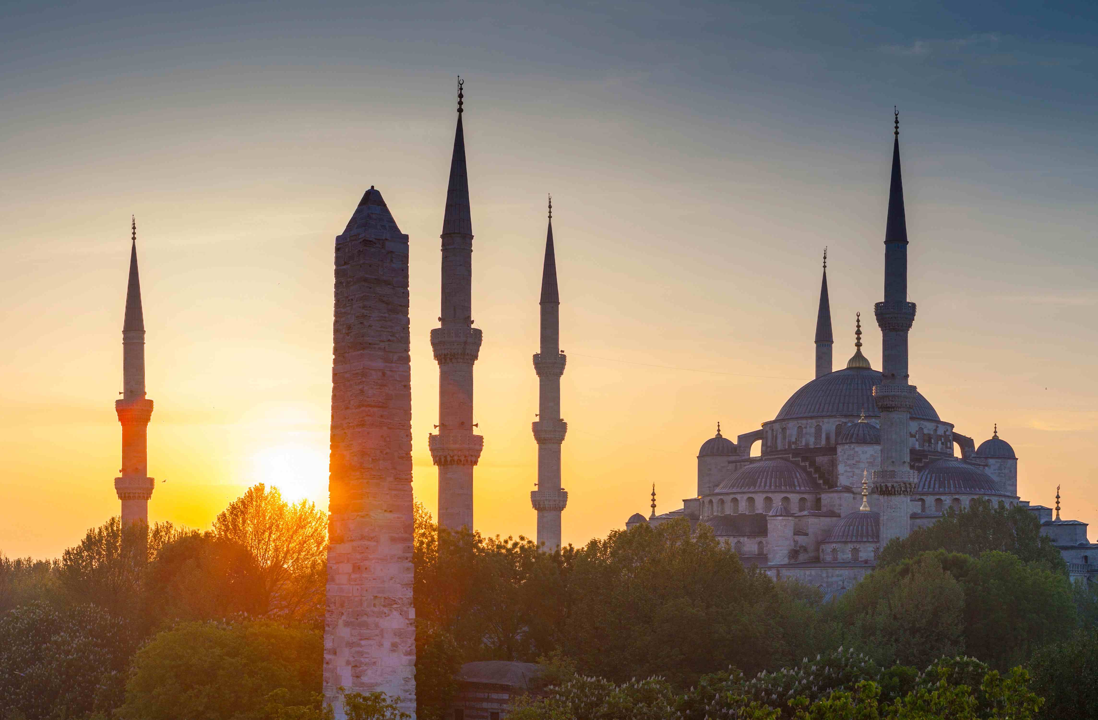 The Blue Mosque (Sultan Ahmed Mosque) in Istanbul at sunset with minarets silhouetted against the sky and trees in the foreground.